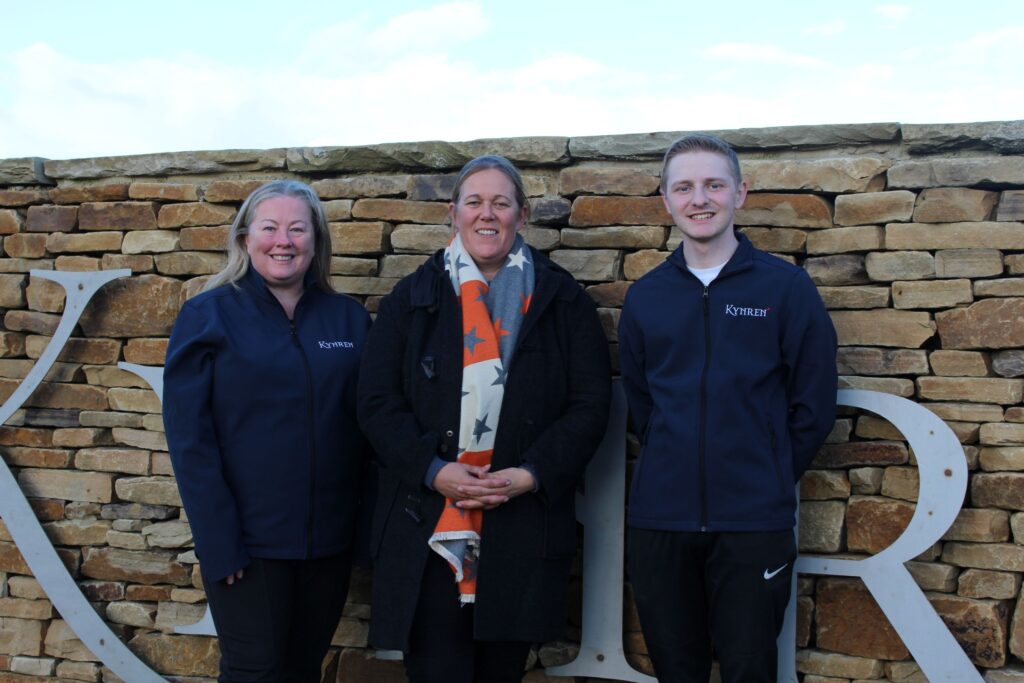 Three Kynren staff members stand smiling to camera in front of a stone Kynren sign