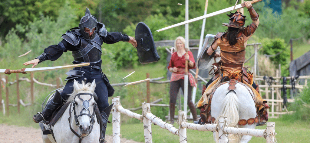 Actors joust on horse back with lances breaking.
