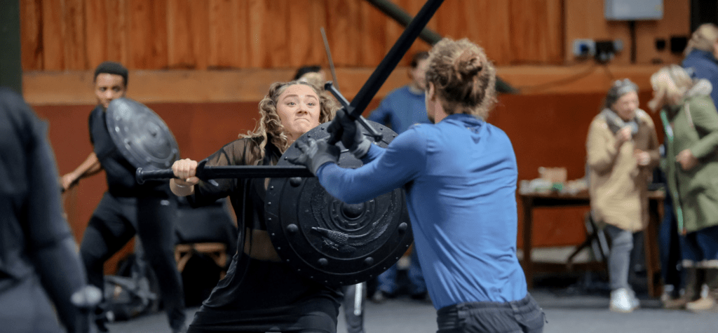 A man and woman holding swords and shields practice combat in an indoor barn
