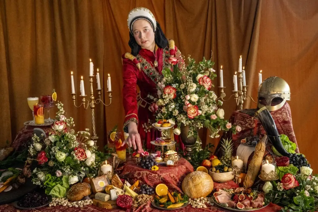 A tudor lady adjusts a macaron on a banquet table laden with food. Kynren, Time Travellers' Feast.