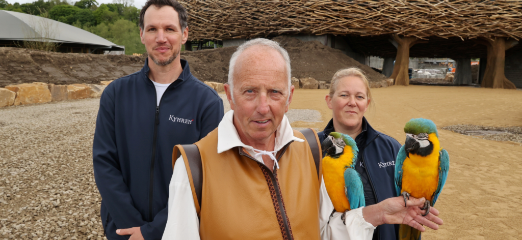 three members of the kynren team, one in costume with 2 macaws on his arm, smile for the camera in front of The Lost Feather arena