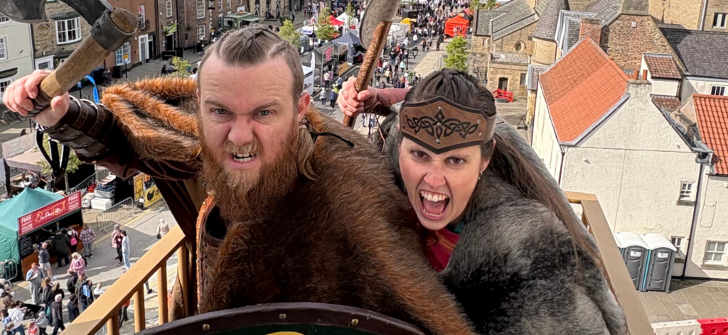 two Vikings on the top of a building shout at the camera with a bustling crowd below.