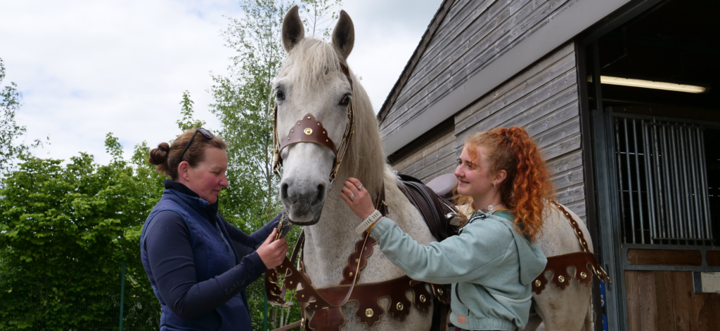 Two women adjust the harness of a horse in front of a block of stables.