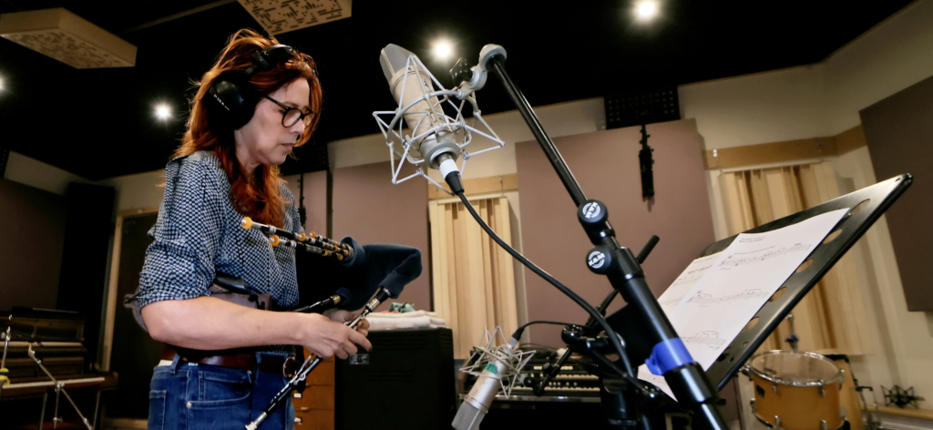 A woman with red hair wearing headphones plays a set of pipes in a recording studio