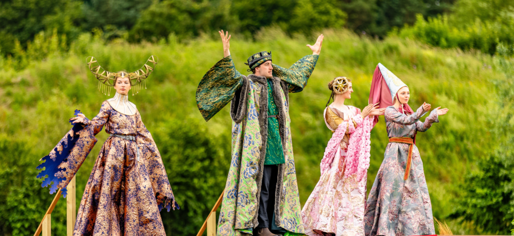 three women and a a man are dressed in ornate medieval-inspired costumes in front of a lush green background