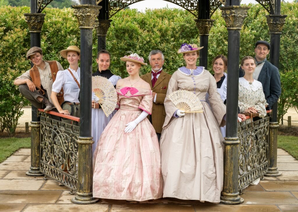 nine characters dressed in various victorian costumes look at the camera from a gazebo in a manicured garden. they all embody their characters as they stare at the viewer