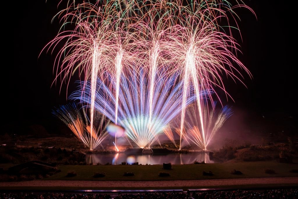 a long exposure shot of fireworks above a huge outdoor stage, complete with a lake