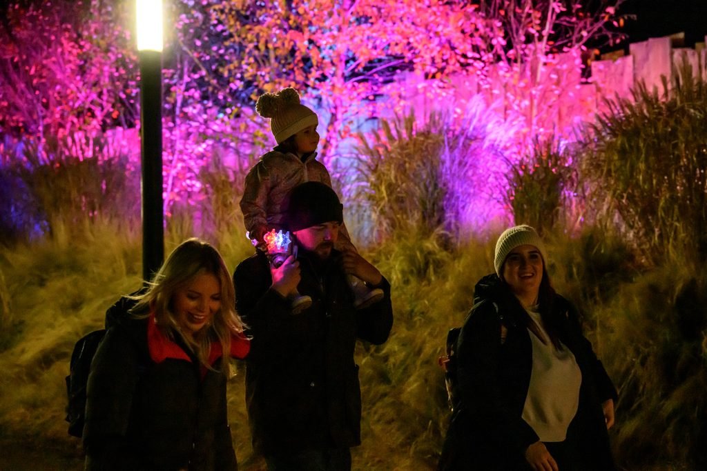 a young girl sits on her father's shoulders as a family walks down a colourfully lit pathway