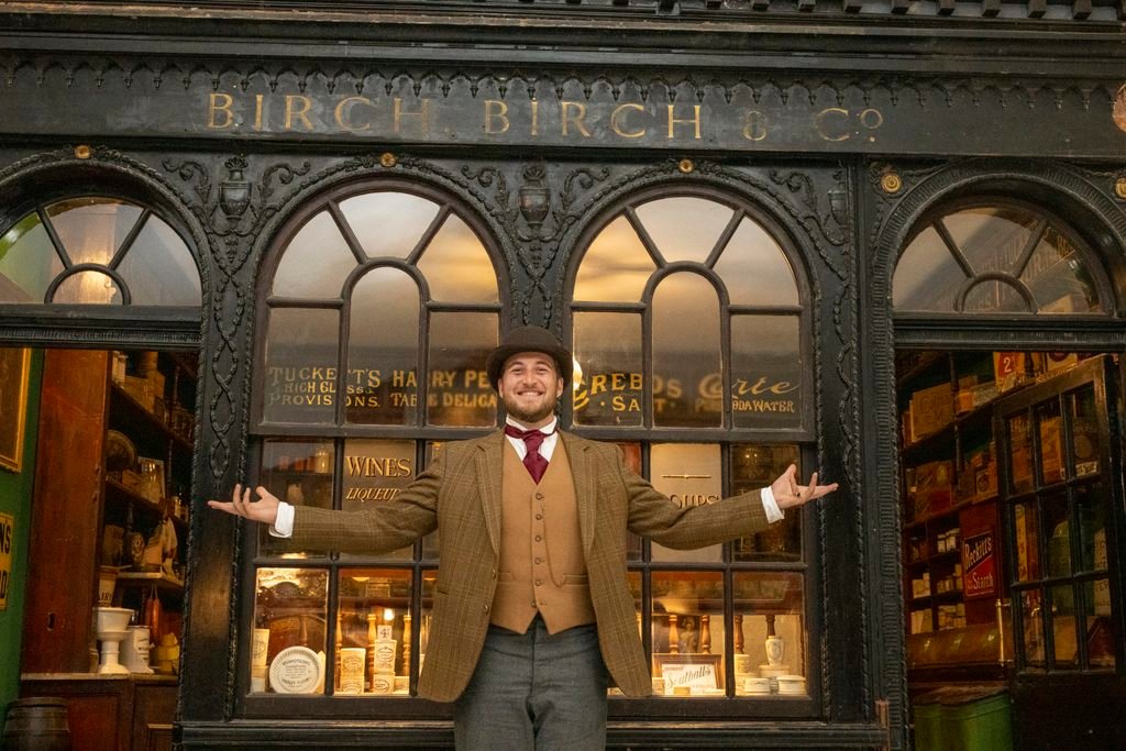 a man dressed in a victorian top hat and suit stands with his arms outstretched in front of an old victorian sweet shop named Birch, Birch & Co
