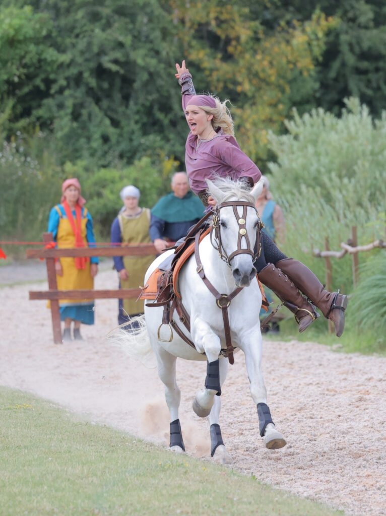 A female trick rider gestures at the crowd mid-stunt as a horse gallops towards the camera.