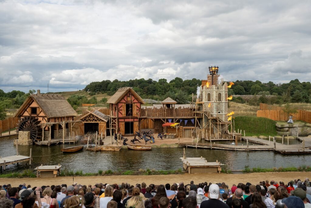 a crowd sits in front of a lakeside medieval village that forms the set of a live-action outdoor show